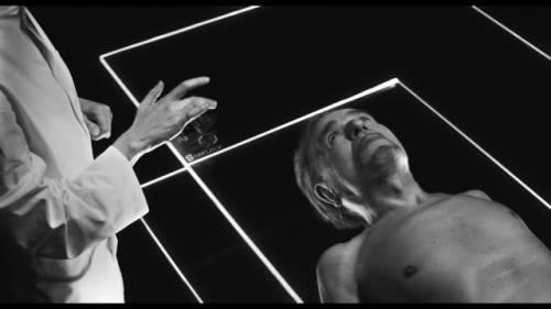 Vignette en noir et blanc extraite du film Panthéon Discount. Un homme, alongé sur une plaque de verre. Au dessus de lui, une seconde plaque de verre de même taille, où les doigts d'une personne en blouse blanche parcourent un écran tactile.